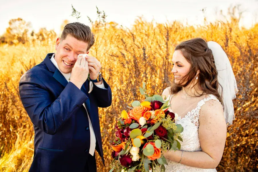 A groom wiping away tears at the first look as his bride holds an autumn bouquet in a golden field