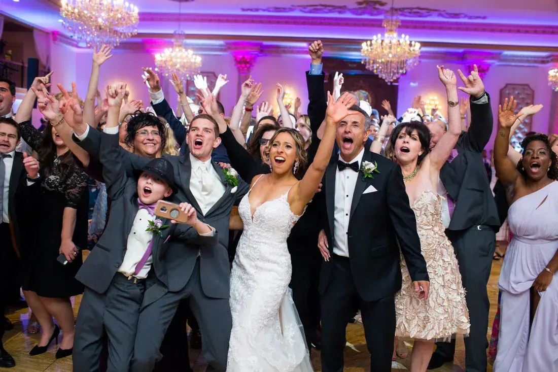 A packed dance floor with the couple and guests raising their arms in a chandelier ballroom