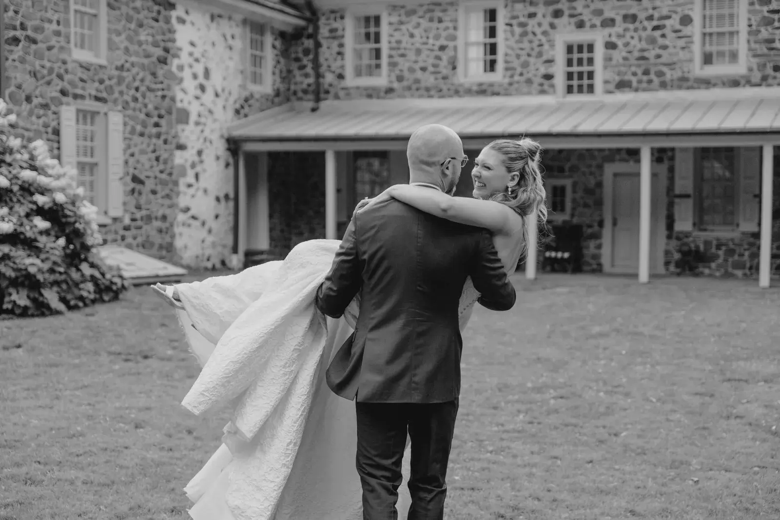 A groom carrying his bride across the lawn of a stone farmhouse, in black and white
