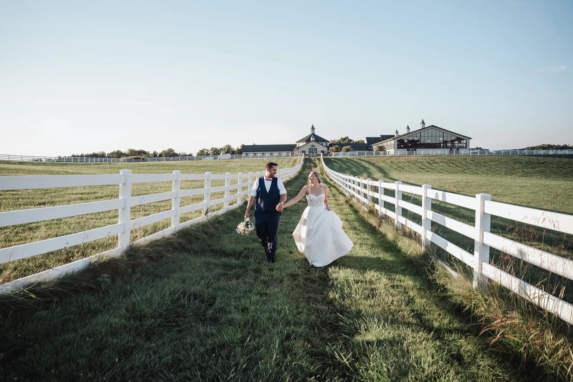 A couple walking hand in hand down a fenced farm lane, photographed by Stephanie Vaccaro